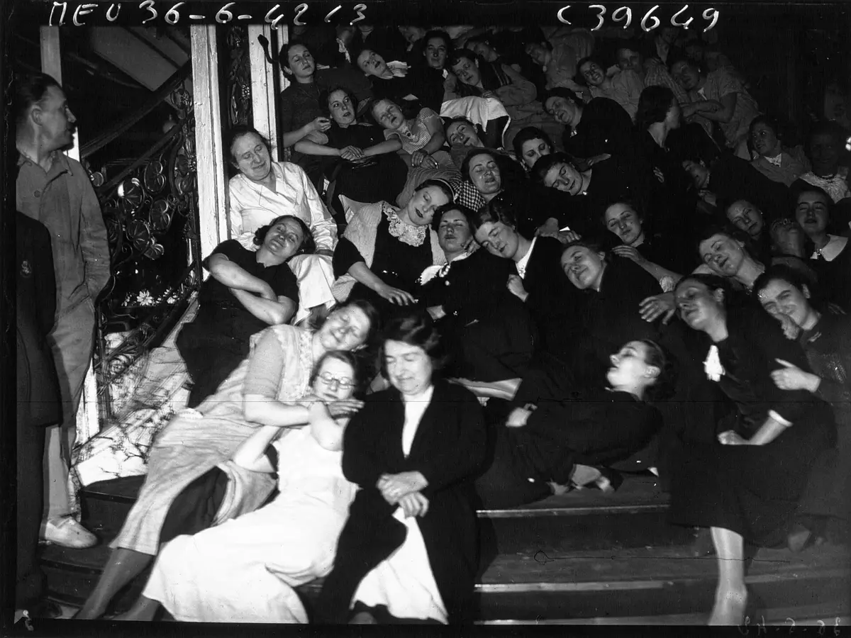 Black-and-white press photo from June 1936: striking department-store employees, mostly women, doze huddled together on a staircase of the store they occupy, some of them smiling.